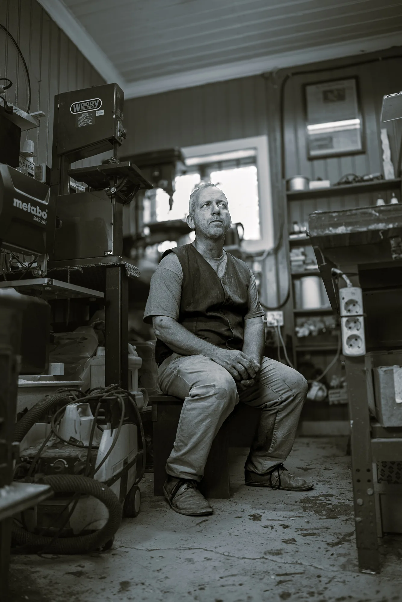 Black and white environmental portrait of a woodworker seated among his machines in a workshop at Ljan Stasjon, Oslo